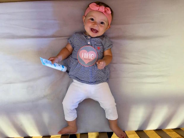 Baby girl nine months old laying in crib and smiling with headband and diaper rash cream