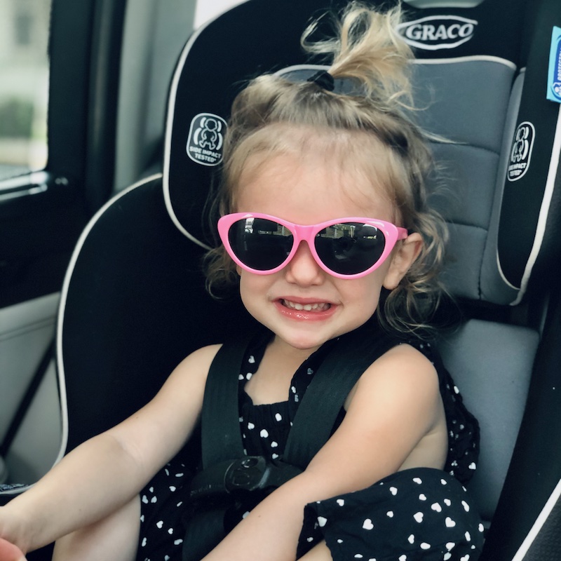 Toddler girl sitting in carseat wearing pink sunglasses and smiling