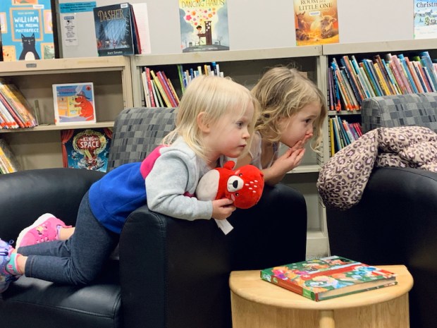 Two toddler girls sitting on a chair at a library while listening to a story