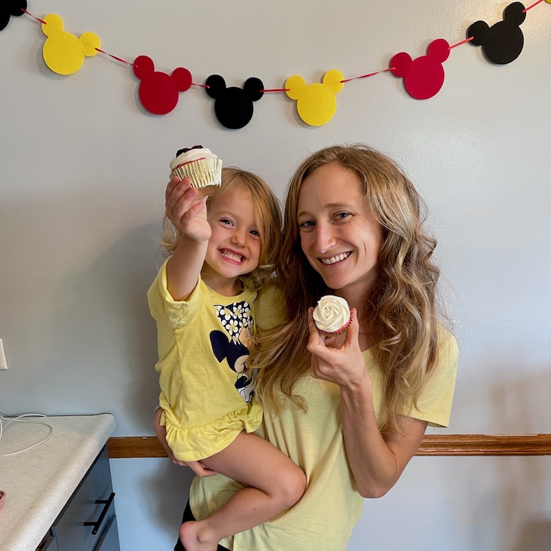 Mom holding toddler and cupcakes with Mickey Mouse birthday decorations