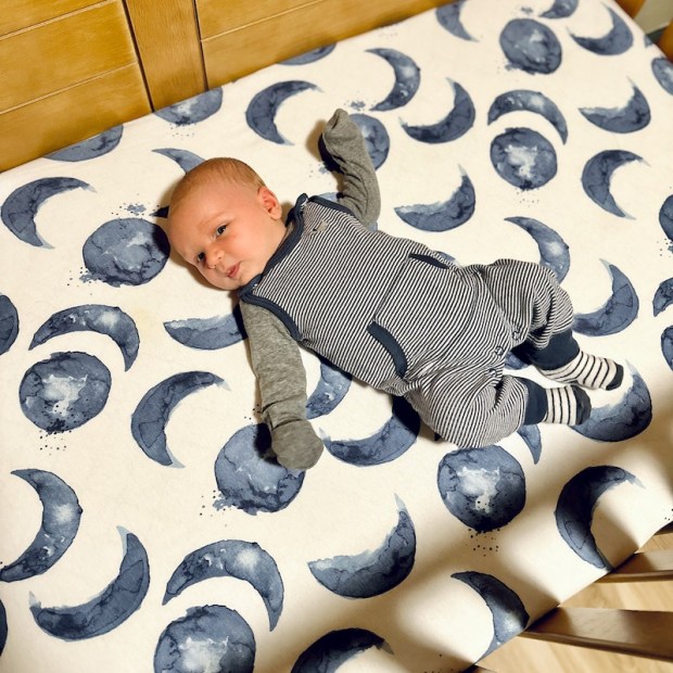 One month old baby boy laying on watercolor moon crib sheet in brown wood crib