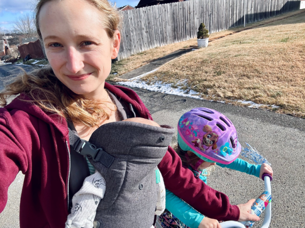 Mom helping daughter ride bike while carrying baby in carrier