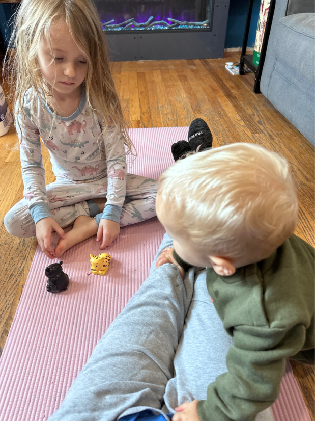 Girl playing with toys while baby crawls on mom sitting on yoga mat