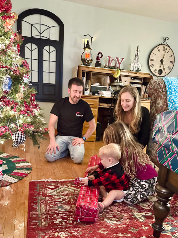 Sister and baby brother opening gifts by Christmas tree with parents sitting nearby