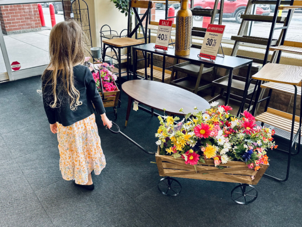 Five year old girl wearing a black cardigan and maxi dress with flowers on it pulling a wagon of fake flowers at Hobby Lobby.