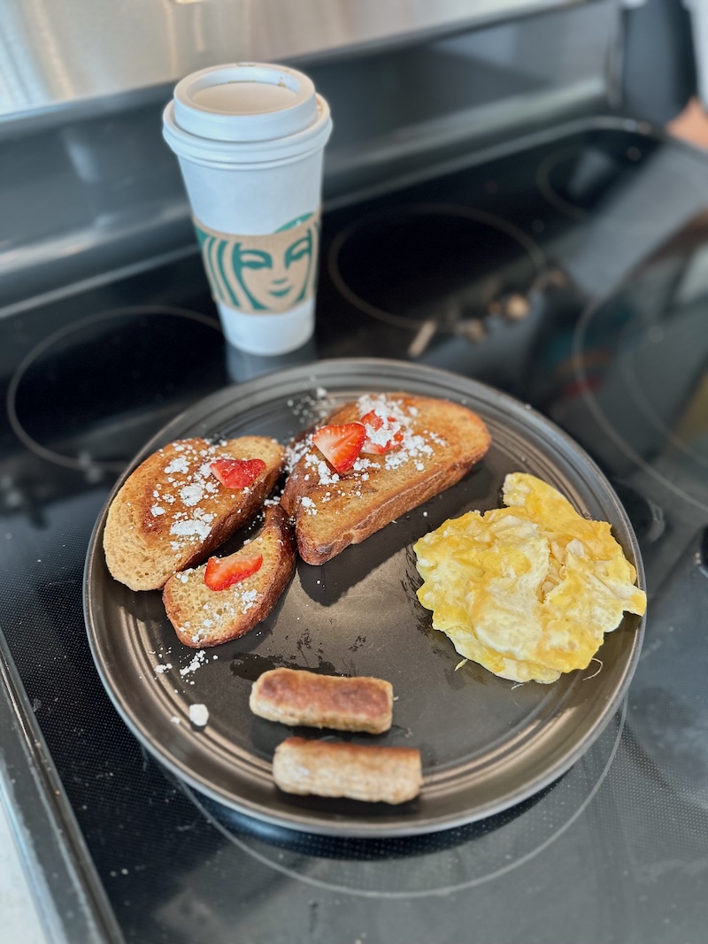 Challah French toast, scrambled eggs, and sausage links on a black plate on top of a stove with Starbucks coffee cup in background