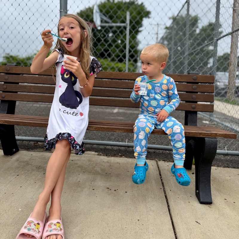 Kids sitting on bench outside of park with yogurt